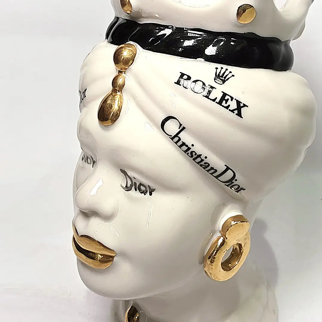 Coppia di Teste di Moro in ceramica bianca lucida con turbanti neri e bianchi decorati da loghi fashion neri e dorati, completate da corone regali e labbra con finitura in oro zecchino. Queste sculture ornamentali di design pop art fondono la tradizione ceramica siciliana con l'estetica del lusso moderno, rappresentando un complemento d'arredo audace ed esclusivo per interni ricercati.
