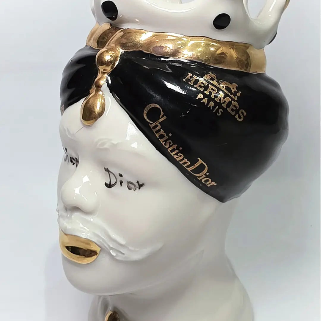 Coppia di Teste di Moro in ceramica bianca lucida con turbanti neri e bianchi decorati da loghi fashion neri e dorati, completate da corone regali e labbra con finitura in oro zecchino. Queste sculture ornamentali di design pop art fondono la tradizione ceramica siciliana con l'estetica del lusso moderno, rappresentando un complemento d'arredo audace ed esclusivo per interni ricercati.
