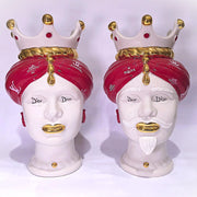 Coppia di Teste di Moro in ceramica bianca lucida con turbanti rosso ciliegia decorati da loghi fashion bianchi, completate da corone regali e labbra con finitura in oro zecchino. Queste sculture ornamentali di design pop art fondono la tradizione ceramica siciliana con l'estetica del lusso moderno, rappresentando un complemento d'arredo audace ed esclusivo per interni ricercati.