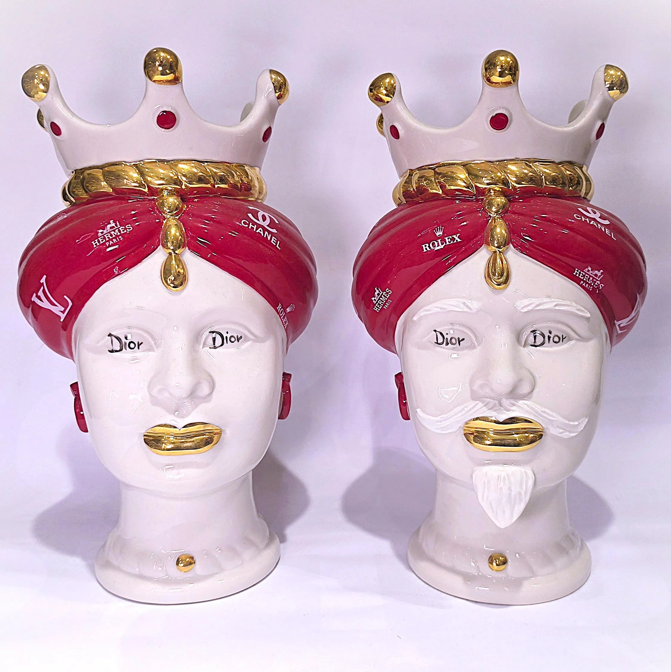 Coppia di Teste di Moro in ceramica bianca lucida con turbanti rosso ciliegia decorati da loghi fashion bianchi, completate da corone regali e labbra con finitura in oro zecchino. Queste sculture ornamentali di design pop art fondono la tradizione ceramica siciliana con l'estetica del lusso moderno, rappresentando un complemento d'arredo audace ed esclusivo per interni ricercati.