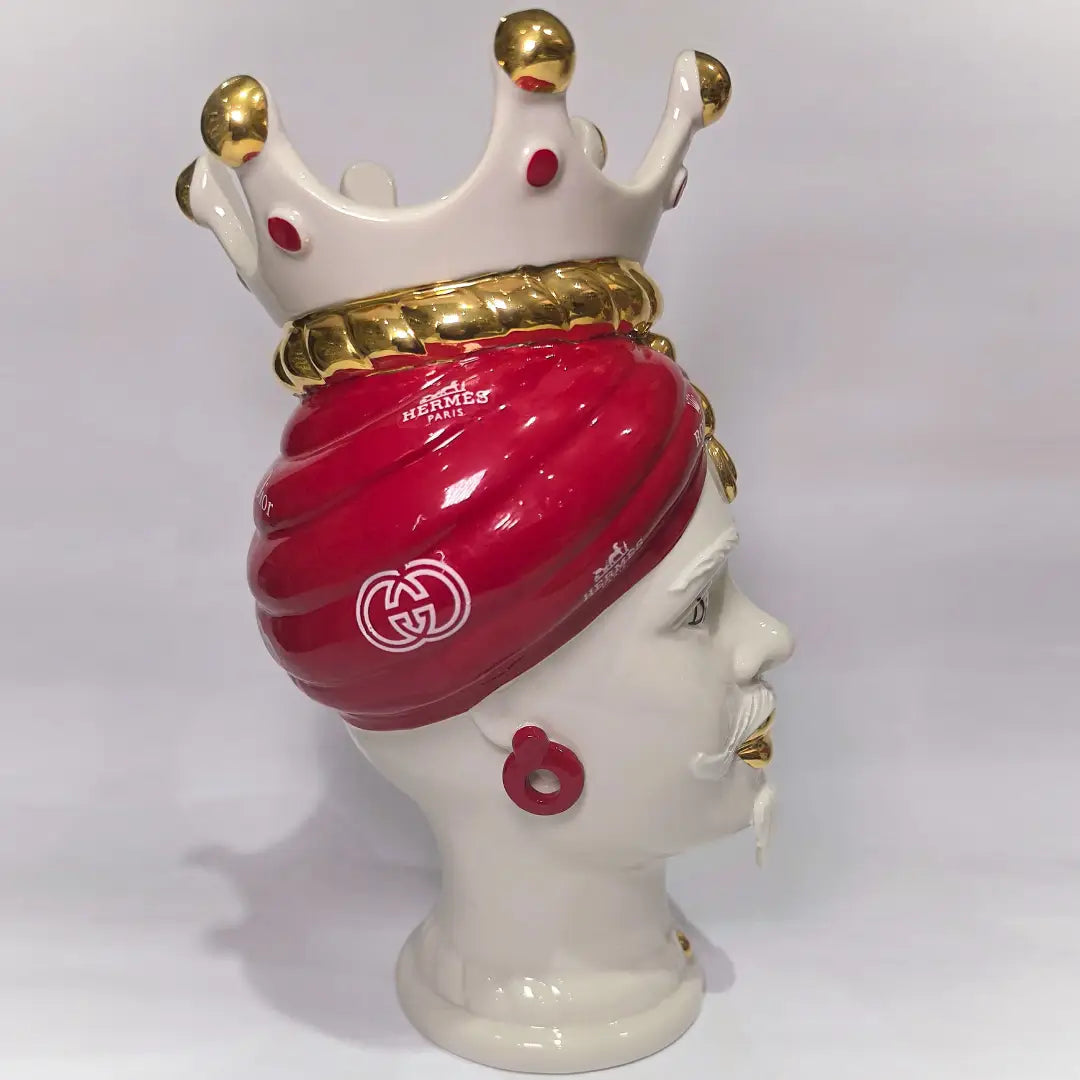 Coppia di Teste di Moro in ceramica bianca lucida con turbanti rosso ciliegia decorati da loghi fashion bianchi, completate da corone regali e labbra con finitura in oro zecchino. Queste sculture ornamentali di design pop art fondono la tradizione ceramica siciliana con l'estetica del lusso moderno, rappresentando un complemento d'arredo audace ed esclusivo per interni ricercati.