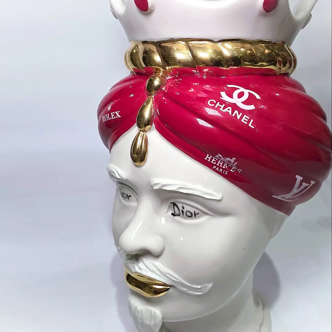 Coppia di Teste di Moro in ceramica bianca lucida con turbanti rosso ciliegia decorati da loghi fashion bianchi, completate da corone regali e labbra con finitura in oro zecchino. Queste sculture ornamentali di design pop art fondono la tradizione ceramica siciliana con l'estetica del lusso moderno, rappresentando un complemento d'arredo audace ed esclusivo per interni ricercati.
