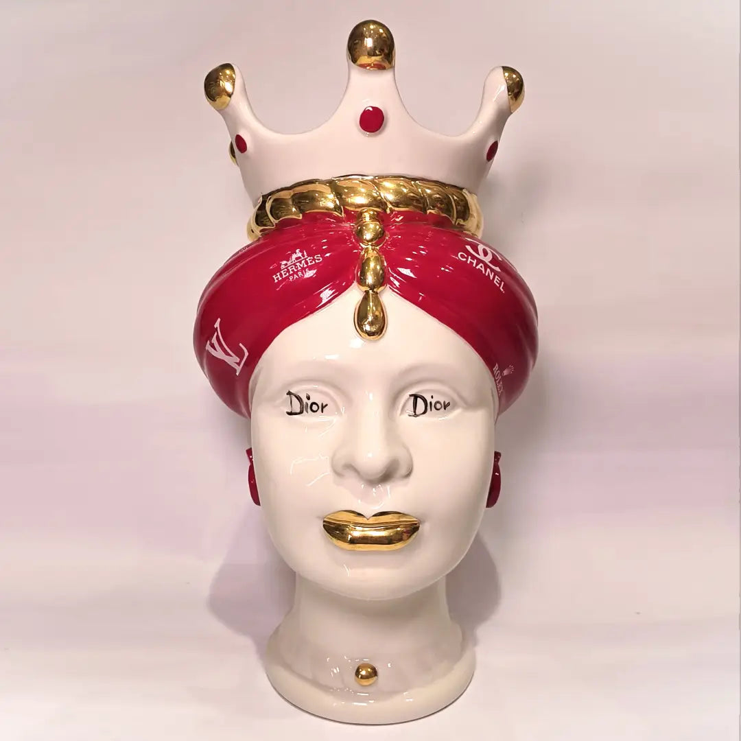 Coppia di Teste di Moro in ceramica bianca lucida con turbanti rosso ciliegia decorati da loghi fashion bianchi, completate da corone regali e labbra con finitura in oro zecchino. Queste sculture ornamentali di design pop art fondono la tradizione ceramica siciliana con l'estetica del lusso moderno, rappresentando un complemento d'arredo audace ed esclusivo per interni ricercati.