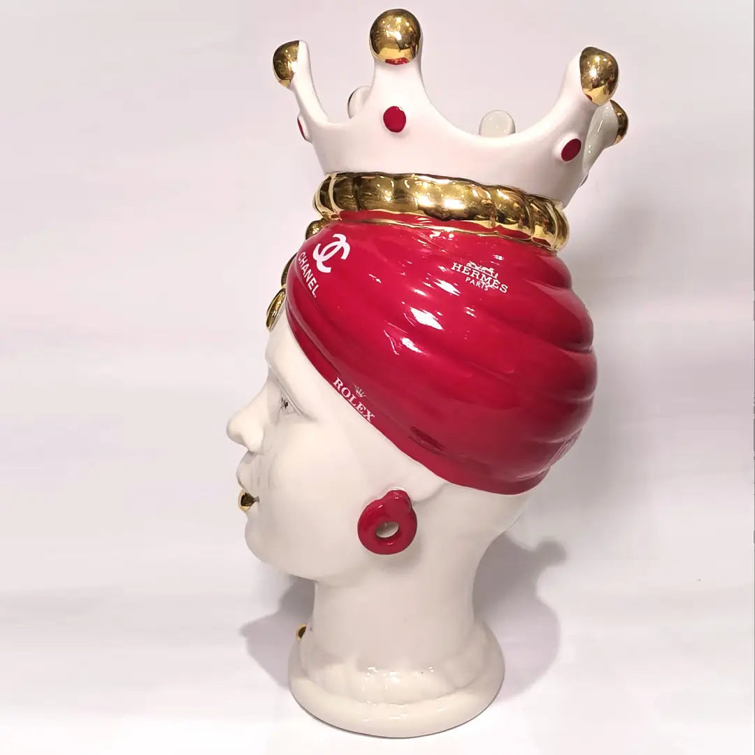 Coppia di Teste di Moro in ceramica bianca lucida con turbanti rosso ciliegia decorati da loghi fashion bianchi, completate da corone regali e labbra con finitura in oro zecchino. Queste sculture ornamentali di design pop art fondono la tradizione ceramica siciliana con l'estetica del lusso moderno, rappresentando un complemento d'arredo audace ed esclusivo per interni ricercati.
