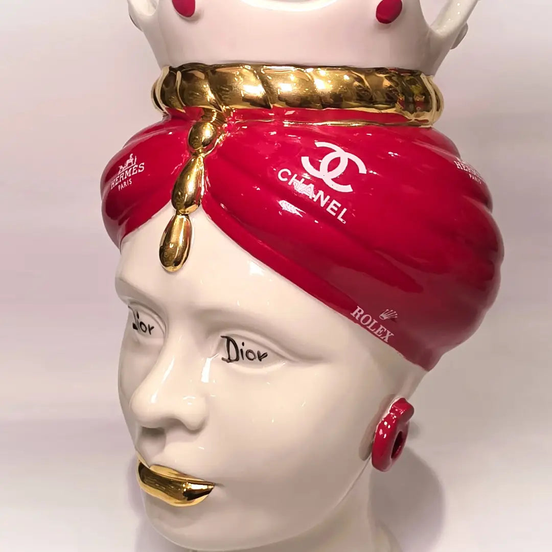Coppia di Teste di Moro in ceramica bianca lucida con turbanti rosso ciliegia decorati da loghi fashion bianchi, completate da corone regali e labbra con finitura in oro zecchino. Queste sculture ornamentali di design pop art fondono la tradizione ceramica siciliana con l'estetica del lusso moderno, rappresentando un complemento d'arredo audace ed esclusivo per interni ricercati.