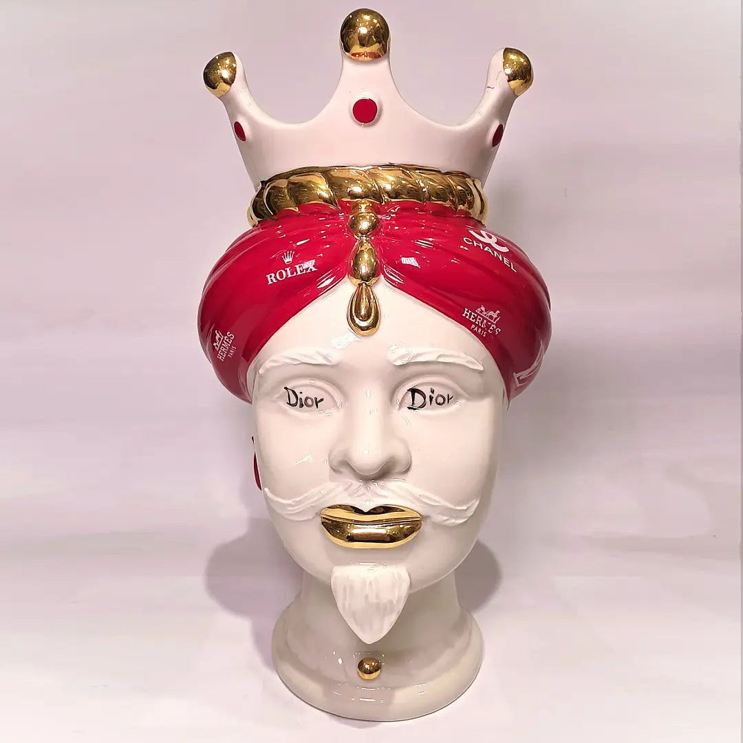 Coppia di Teste di Moro in ceramica bianca lucida con turbanti rosso ciliegia decorati da loghi fashion bianchi, completate da corone regali e labbra con finitura in oro zecchino. Queste sculture ornamentali di design pop art fondono la tradizione ceramica siciliana con l'estetica del lusso moderno, rappresentando un complemento d'arredo audace ed esclusivo per interni ricercati.
