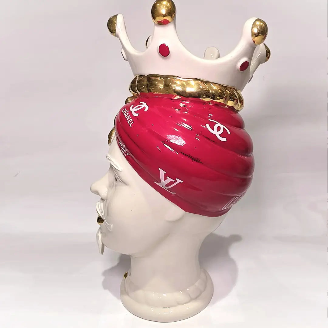 Coppia di Teste di Moro in ceramica bianca lucida con turbanti rosso ciliegia decorati da loghi fashion bianchi, completate da corone regali e labbra con finitura in oro zecchino. Queste sculture ornamentali di design pop art fondono la tradizione ceramica siciliana con l'estetica del lusso moderno, rappresentando un complemento d'arredo audace ed esclusivo per interni ricercati.