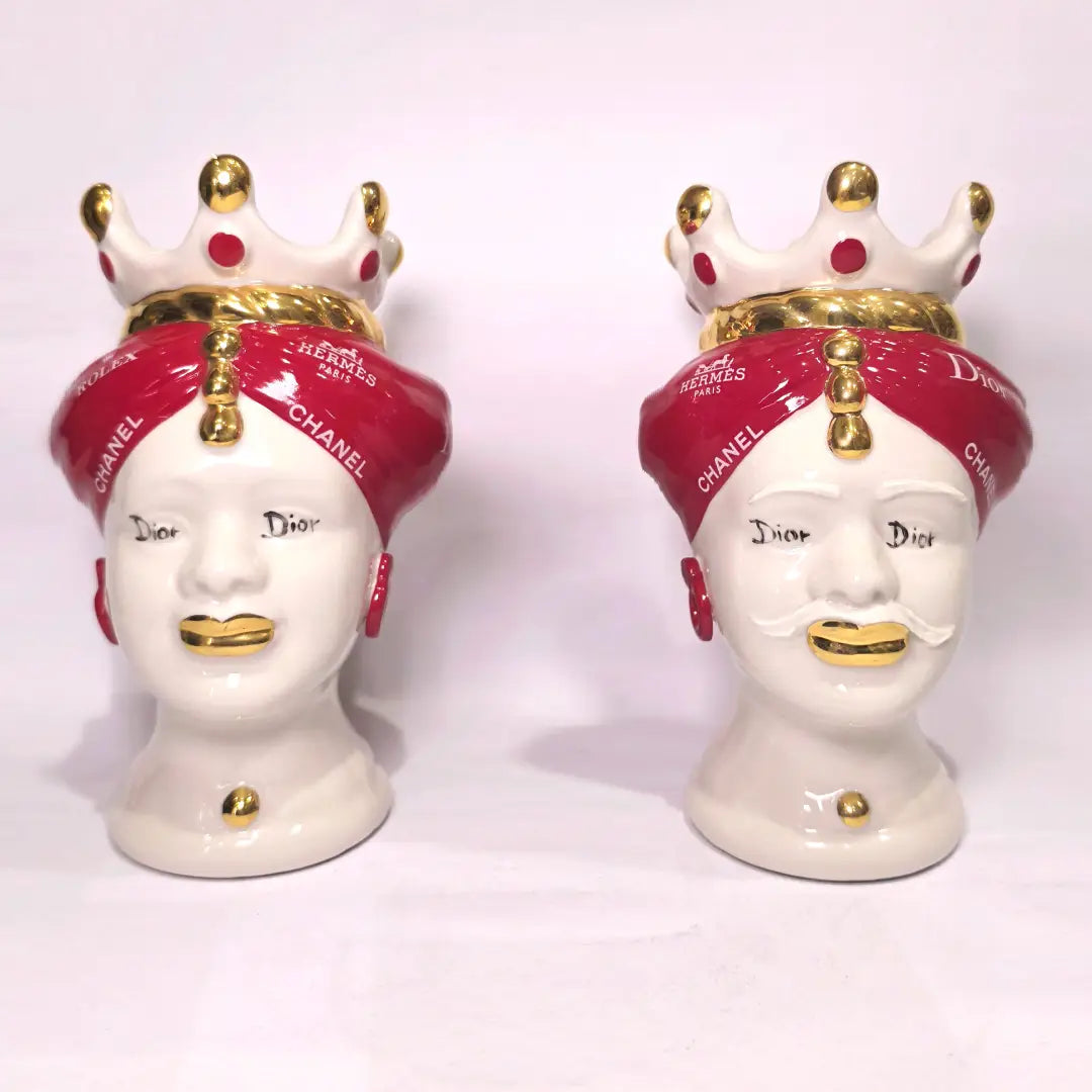 Coppia di Teste di Moro in ceramica bianca lucida con turbanti rosso ciliegia decorati da loghi fashion bianchi, completate da corone regali e labbra con finitura in oro zecchino. Queste sculture ornamentali di design pop art fondono la tradizione ceramica siciliana con l'estetica del lusso moderno, rappresentando un complemento d'arredo audace ed esclusivo per interni ricercati.