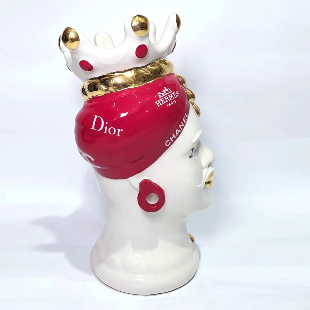 Coppia di Teste di Moro in ceramica bianca lucida con turbanti rosso ciliegia decorati da loghi fashion bianchi, completate da corone regali e labbra con finitura in oro zecchino. Queste sculture ornamentali di design pop art fondono la tradizione ceramica siciliana con l'estetica del lusso moderno, rappresentando un complemento d'arredo audace ed esclusivo per interni ricercati.