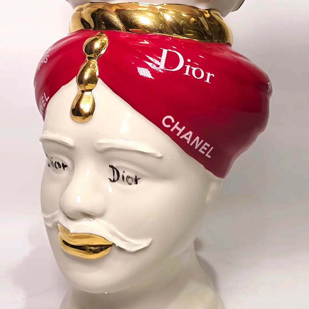 Coppia di Teste di Moro in ceramica bianca lucida con turbanti rosso ciliegia decorati da loghi fashion bianchi, completate da corone regali e labbra con finitura in oro zecchino. Queste sculture ornamentali di design pop art fondono la tradizione ceramica siciliana con l'estetica del lusso moderno, rappresentando un complemento d'arredo audace ed esclusivo per interni ricercati.
