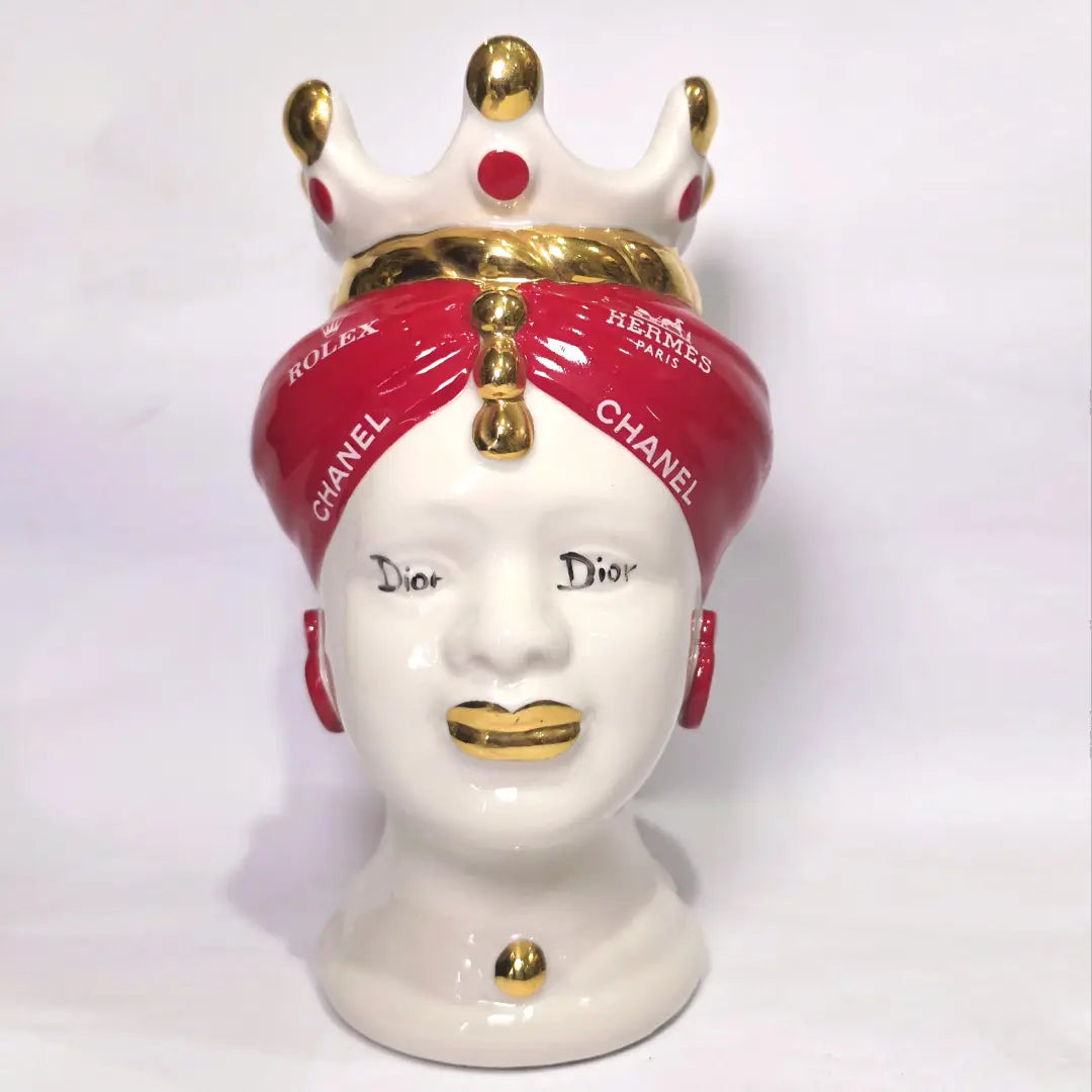 Coppia di Teste di Moro in ceramica bianca lucida con turbanti rosso ciliegia decorati da loghi fashion bianchi, completate da corone regali e labbra con finitura in oro zecchino. Queste sculture ornamentali di design pop art fondono la tradizione ceramica siciliana con l'estetica del lusso moderno, rappresentando un complemento d'arredo audace ed esclusivo per interni ricercati.