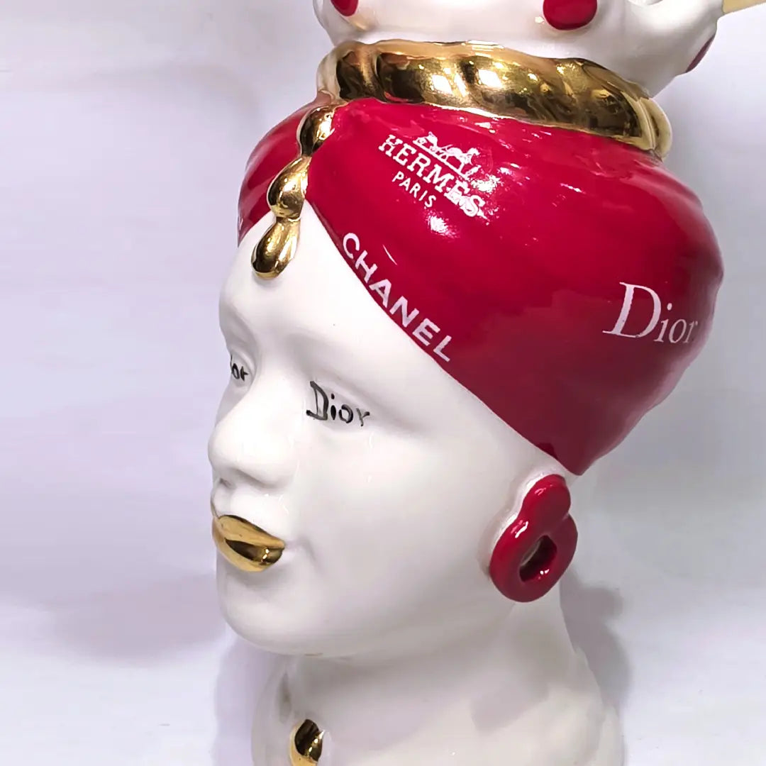 Coppia di Teste di Moro in ceramica bianca lucida con turbanti rosso ciliegia decorati da loghi fashion bianchi, completate da corone regali e labbra con finitura in oro zecchino. Queste sculture ornamentali di design pop art fondono la tradizione ceramica siciliana con l'estetica del lusso moderno, rappresentando un complemento d'arredo audace ed esclusivo per interni ricercati.