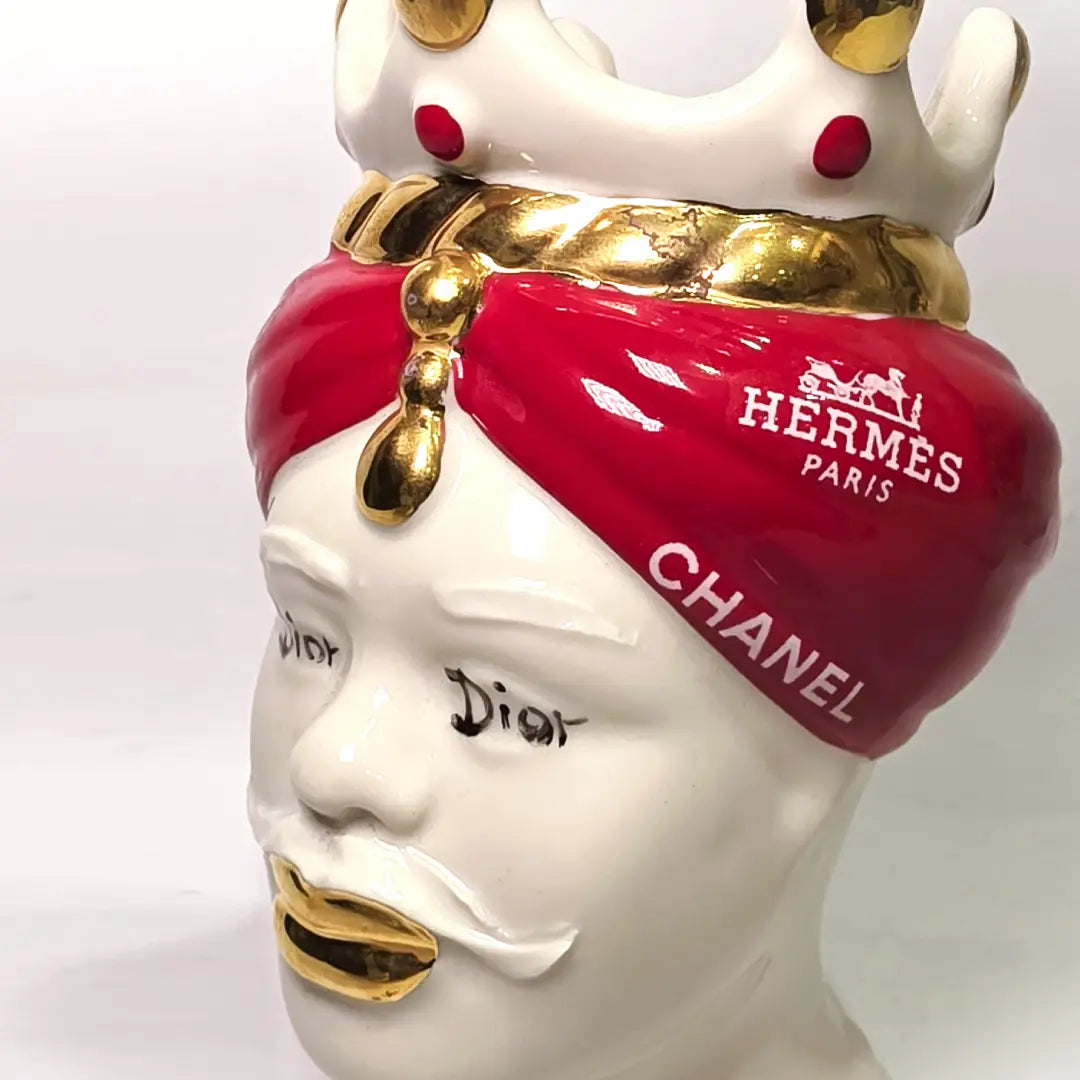 Coppia di Teste di Moro in ceramica bianca lucida con turbanti rosso ciliegia decorati da loghi fashion bianchi, completate da corone regali e labbra con finitura in oro zecchino. Queste sculture ornamentali di design pop art fondono la tradizione ceramica siciliana con l'estetica del lusso moderno, rappresentando un complemento d'arredo audace ed esclusivo per interni ricercati.