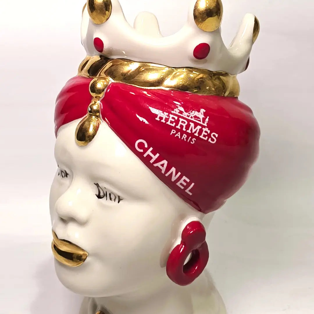 Coppia di Teste di Moro in ceramica bianca lucida con turbanti rosso ciliegia decorati da loghi fashion bianchi, completate da corone regali e labbra con finitura in oro zecchino. Queste sculture ornamentali di design pop art fondono la tradizione ceramica siciliana con l'estetica del lusso moderno, rappresentando un complemento d'arredo audace ed esclusivo per interni ricercati.