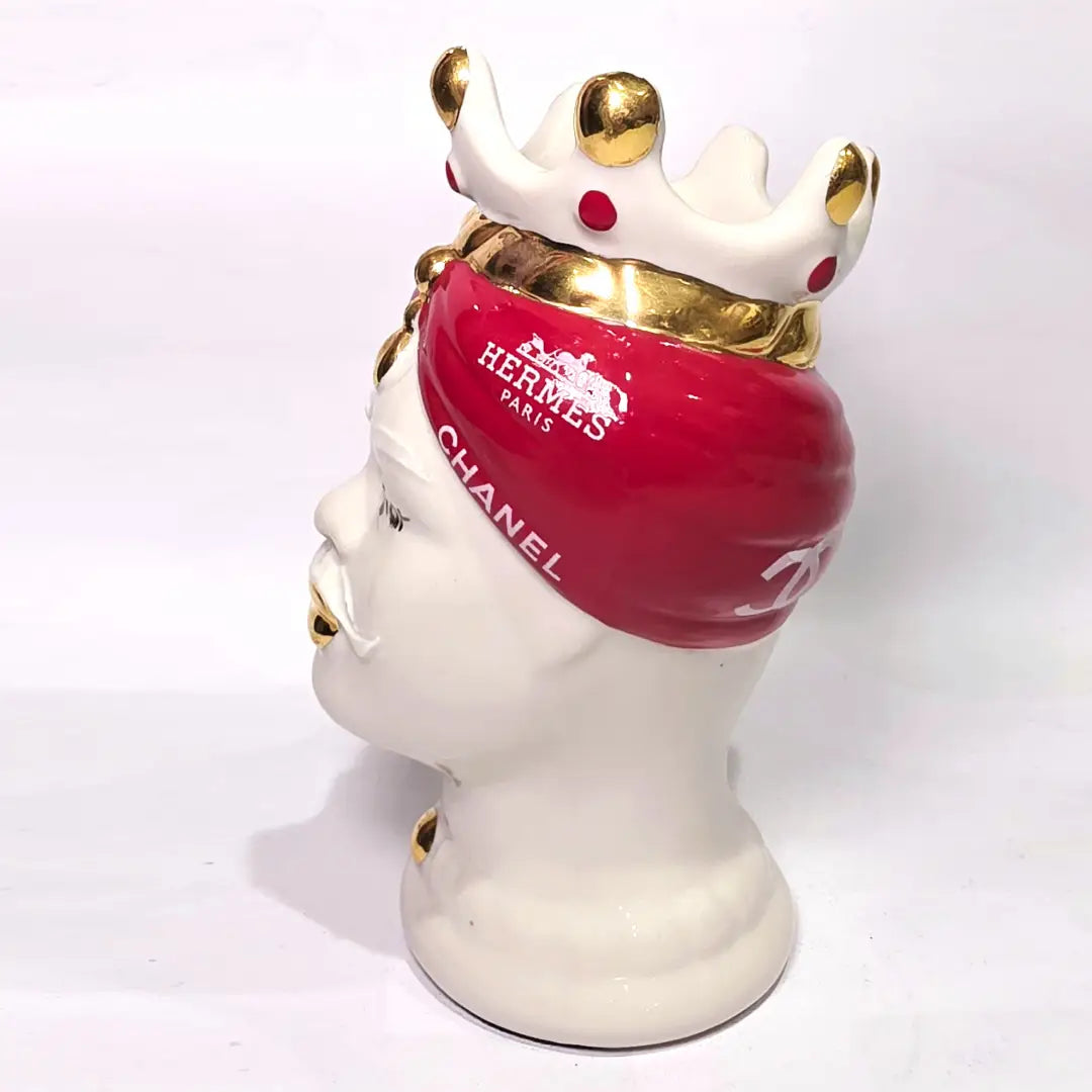 Coppia di Teste di Moro in ceramica bianca lucida con turbanti rosso ciliegia decorati da loghi fashion bianchi, completate da corone regali e labbra con finitura in oro zecchino. Queste sculture ornamentali di design pop art fondono la tradizione ceramica siciliana con l'estetica del lusso moderno, rappresentando un complemento d'arredo audace ed esclusivo per interni ricercati.