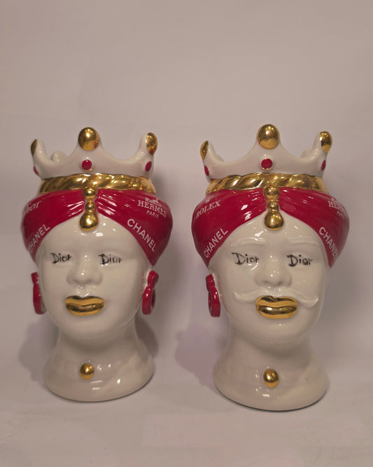 Coppia di Teste di Moro in ceramica bianca lucida con turbanti rosso ciliegia decorati da loghi fashion bianchi, completate da corone regali e labbra con finitura in oro zecchino. Queste sculture ornamentali di design pop art fondono la tradizione ceramica siciliana con l'estetica del lusso moderno, rappresentando un complemento d'arredo audace ed esclusivo per interni ricercati.