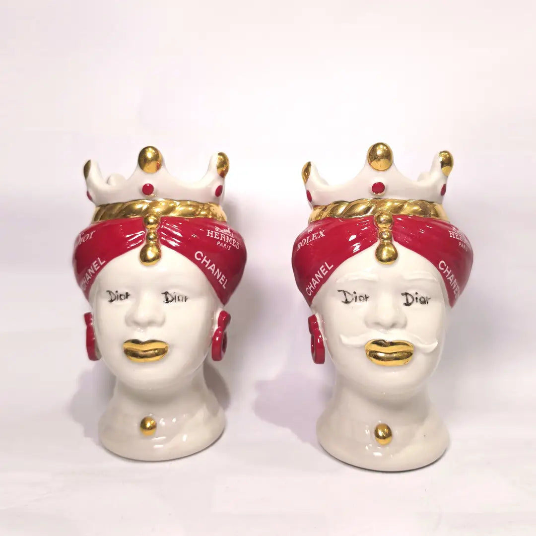 Coppia di Teste di Moro in ceramica bianca lucida con turbanti rosso ciliegia decorati da loghi fashion bianchi, completate da corone regali e labbra con finitura in oro zecchino. Queste sculture ornamentali di design pop art fondono la tradizione ceramica siciliana con l'estetica del lusso moderno, rappresentando un complemento d'arredo audace ed esclusivo per interni ricercati.