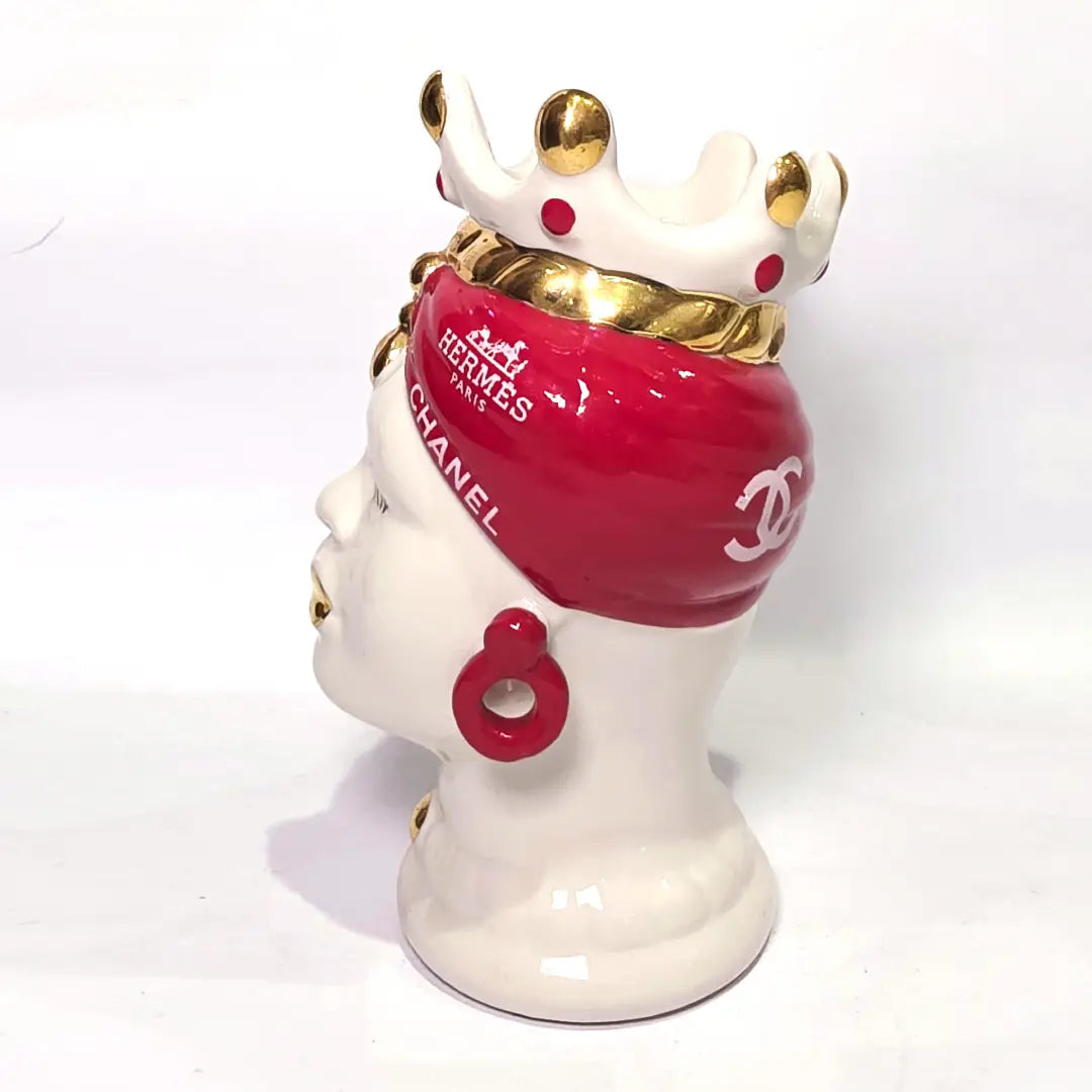Coppia di Teste di Moro in ceramica bianca lucida con turbanti rosso ciliegia decorati da loghi fashion bianchi, completate da corone regali e labbra con finitura in oro zecchino. Queste sculture ornamentali di design pop art fondono la tradizione ceramica siciliana con l'estetica del lusso moderno, rappresentando un complemento d'arredo audace ed esclusivo per interni ricercati.