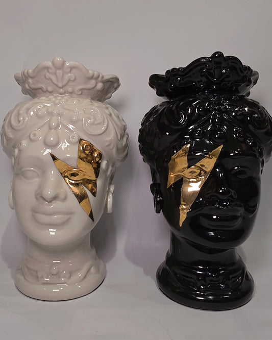 Coppia di Teste di Moro in ceramica artigianale, una in bianco lucido e una in nero lucido, entrambe caratterizzate da un'originale saetta dorata dipinta sul volto. Queste sculture ornamentali dal design pop e contemporaneo fondono la tradizione siciliana con uno stile d'avanguardia, rappresentando un complemento d'arredo iconico ed elegante per interni di lusso.