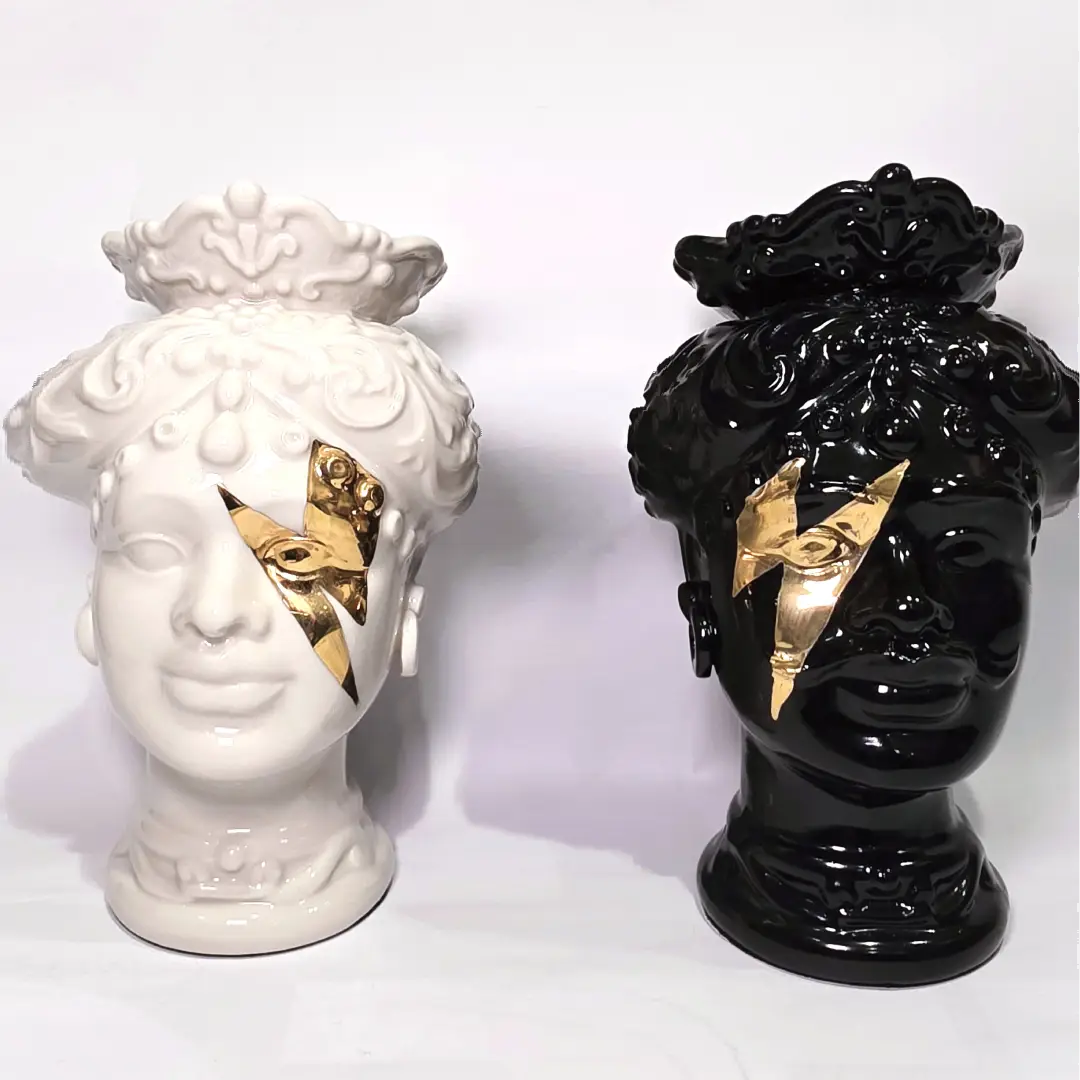 Coppia di Teste di Moro in ceramica artigianale, una in bianco lucido e una in nero lucido, entrambe caratterizzate da un'originale saetta dorata dipinta sul volto. Queste sculture ornamentali dal design pop e contemporaneo fondono la tradizione siciliana con uno stile d'avanguardia, rappresentando un complemento d'arredo iconico ed elegante per interni di lusso.