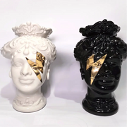 Coppia di Teste di Moro in ceramica artigianale, una in bianco lucido e una in nero lucido, entrambe caratterizzate da un'originale saetta dorata dipinta sul volto. Queste sculture ornamentali dal design pop e contemporaneo fondono la tradizione siciliana con uno stile d'avanguardia, rappresentando un complemento d'arredo iconico ed elegante per interni di lusso.