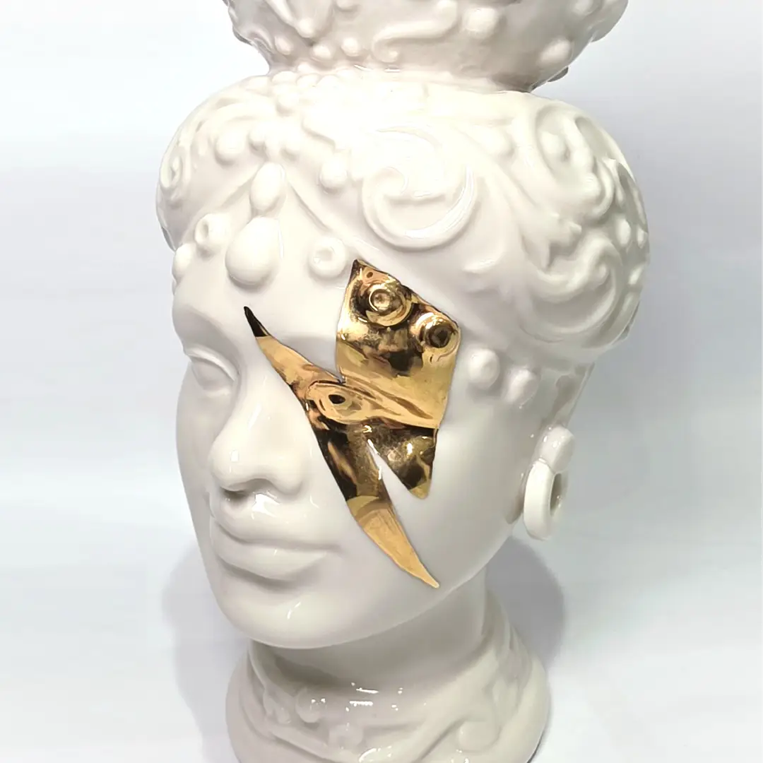 Coppia di Teste di Moro in ceramica artigianale, una in bianco lucido e una in nero lucido, entrambe caratterizzate da un'originale saetta dorata dipinta sul volto. Queste sculture ornamentali dal design pop e contemporaneo fondono la tradizione siciliana con uno stile d'avanguardia, rappresentando un complemento d'arredo iconico ed elegante per interni di lusso.