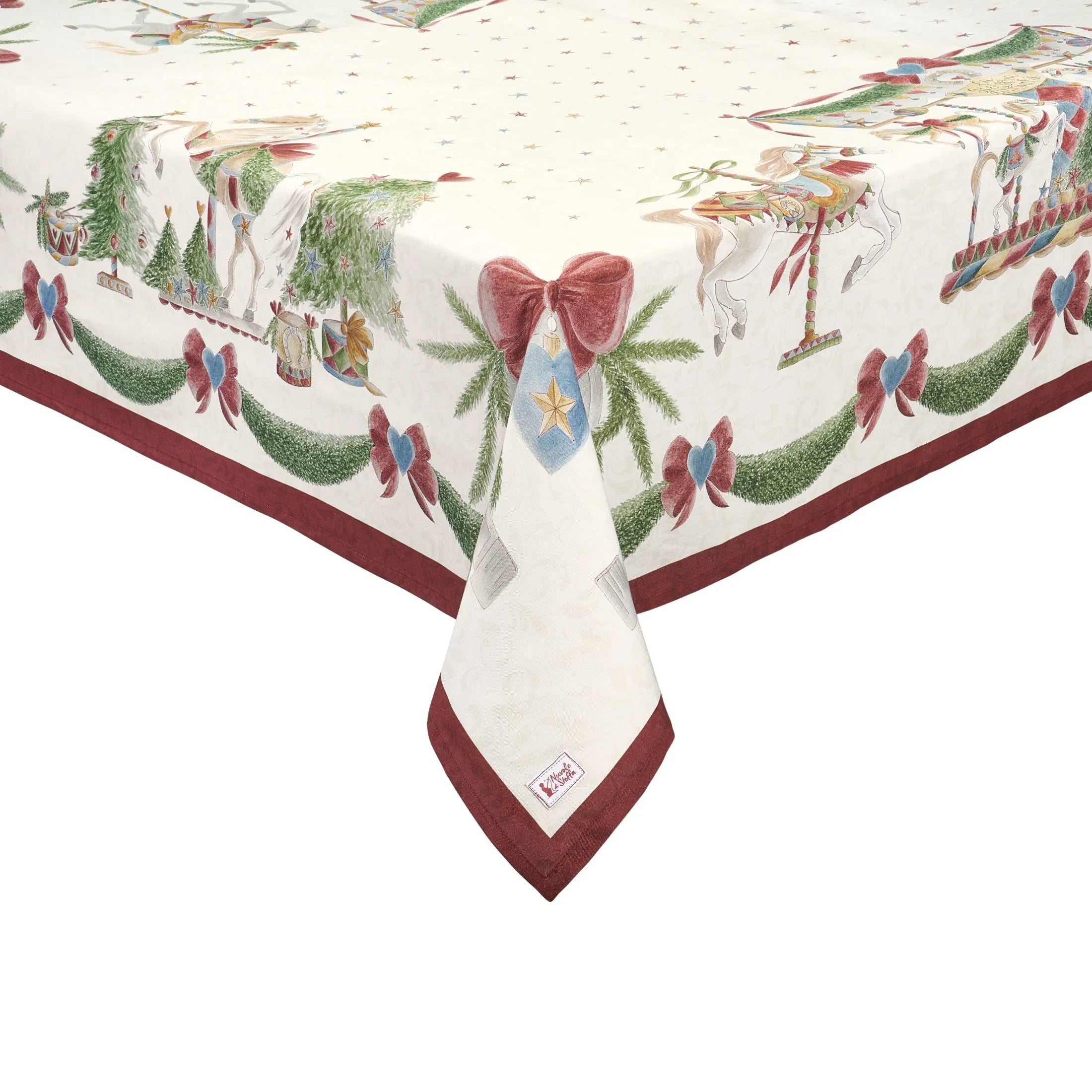 Dettaglio dell'angolo di una tovaglia natalizia in cotone Nuvole di Stoffa "La Giostra di Amelie". Il design presenta cavalli da giostra, ghirlande di pino e fiocchi rossi su un fondo panna stellato, rifinito da un bordo rosso scuro e l'etichetta del brand sull'orlo.