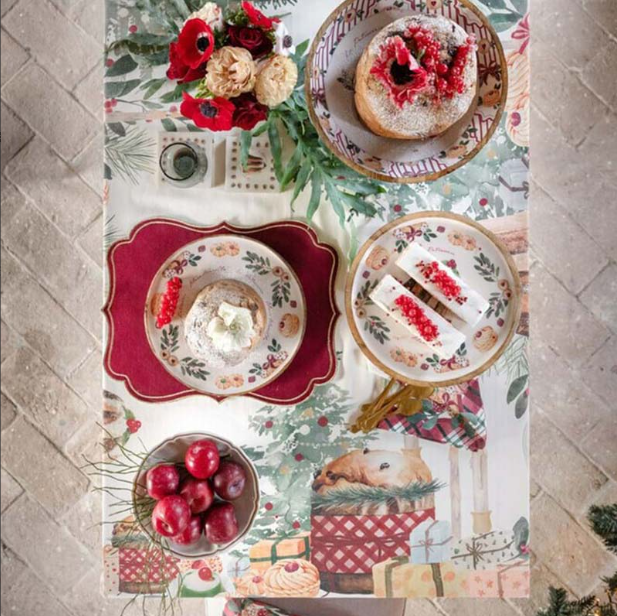 Inquadratura dall'alto di una tavola imbandita con tovaglia Blanc Mariclo. Sono visibili piatti decorati con panettoni e ribes, una ciotola di mele rosse, un mazzo di anemoni e decorazioni natalizie.