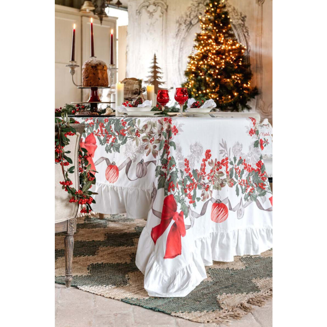Tavola imbandita con tovaglia a gala Sinfonia Natalizia. Sul piano, calici rossi, candele e panettone. Sullo sfondo un albero di Natale illuminato e pareti decorate creano un'atmosfera calda ed elegante.