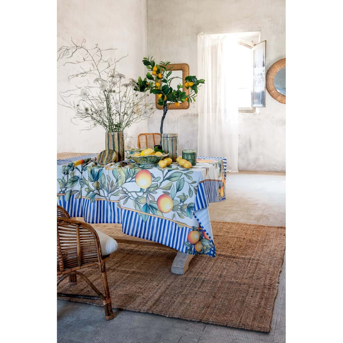 Ambientazione tovaglia Blanc Mariclo' linea Ortigia con stampa a righe blu. Decoro mediterraneo con rami di limoni e motivi ornamentali. Tessuto d'arredo elegante per tavola estiva.