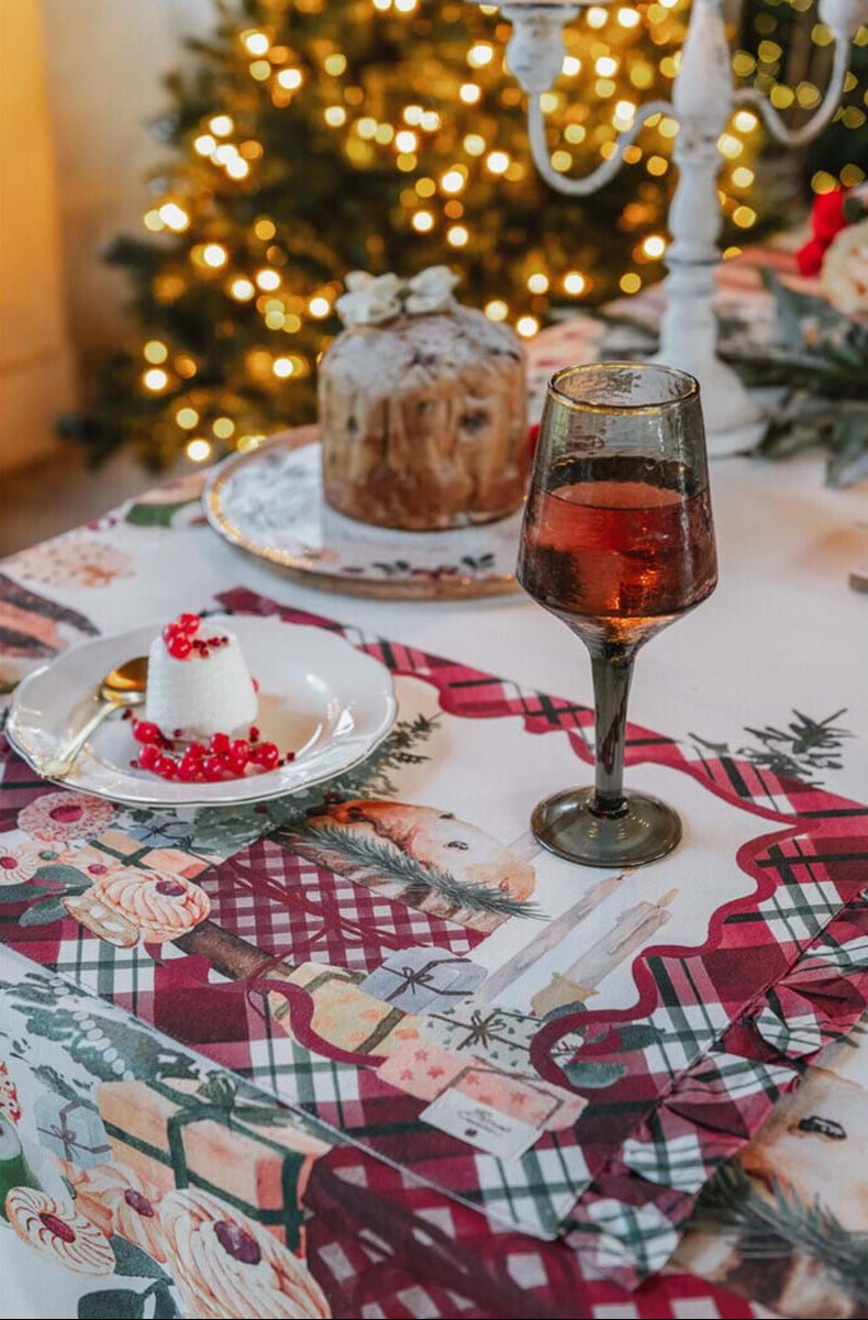 Dettaglio mise en place natalizia Blanc Mariclo con tovaglietta americana a balze. Sopra, un calice di vino, un piattino con dessert e ribes, e un panettone, con albero di Natale illuminato sfocato.