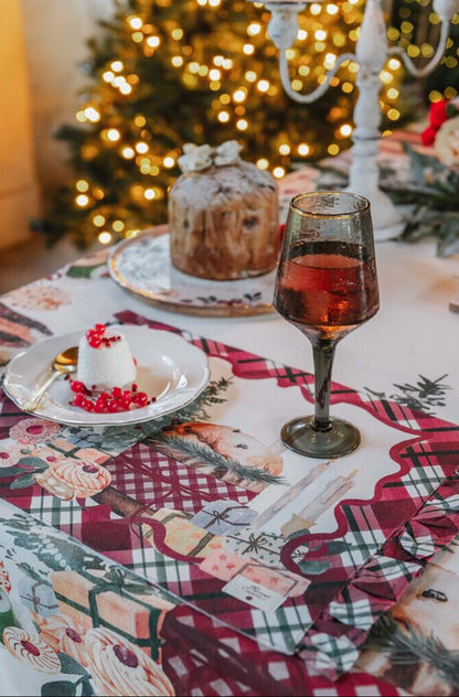 Dettaglio mise en place natalizia Blanc Mariclo con tovaglietta americana a balze. Sopra, un calice di vino, un piattino con dessert e ribes, e un panettone, con albero di Natale illuminato sfocato.