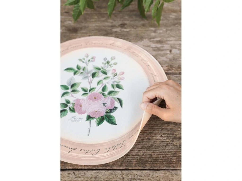 Primo piano di una mano che solleva leggermente il bordo di una tovaglietta tonda in vinile "Le Botanique" di Blanc Mariclò, mostrandone la flessibilità. La tovaglietta è appoggiata su un tavolo di legno rustico e presenta una stampa di rose rosa.