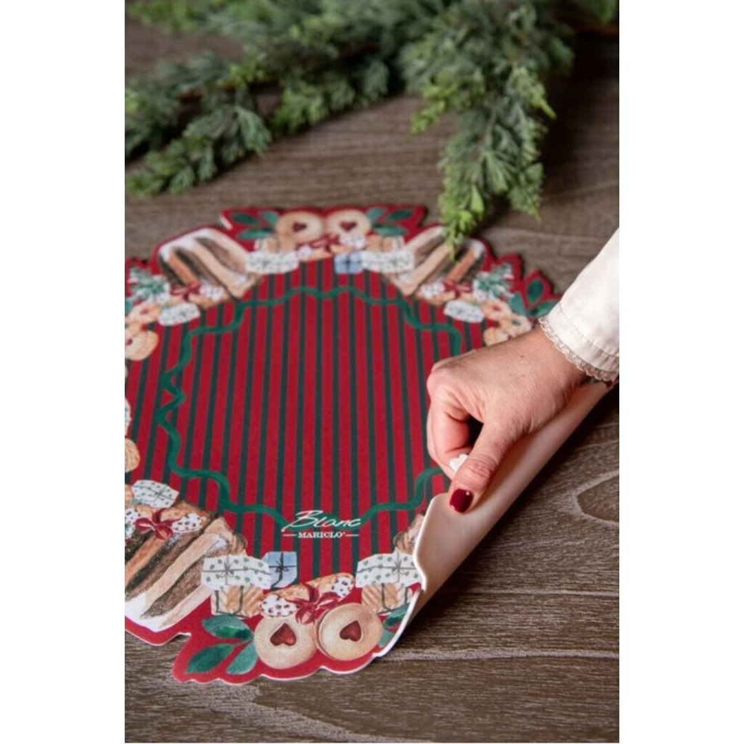 Dettaglio di una tovaglietta in vinile Blanc Mariclo sollevata da una mano per mostrarne la flessibilità. Il design tondo presenta righe rosse e verdi centrali e una ghirlanda di dolci natalizi.