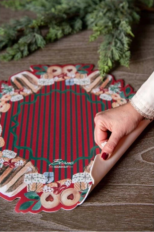 Dettaglio di una tovaglietta in vinile Blanc Mariclo sollevata da una mano per mostrarne la flessibilità. Il design tondo presenta righe rosse e verdi centrali e una ghirlanda di dolci natalizi.