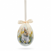 Decorazione pasquale Blanc Mariclò a forma di uovo giallo paglierino con nastro in raso crema. Il decoro centrale raffigura un coniglietto bianco seduto tra narcisi gialli e piccoli fiori bianchi di campo.