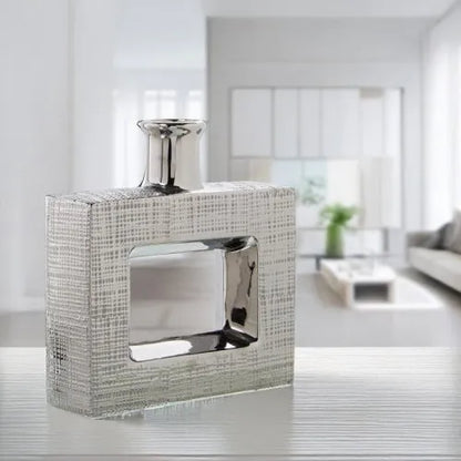 Vaso a bottiglia rettangolare orizzontale firmato e-Camacho, caratterizzato da un'elegante finitura ceramica color argento lucido con una texture a rilievo finemente lavorata e un originale design geometrico con foro centrale vuoto. Questo moderno complemento d'arredo è provvisto di un collo stretto superiore ideale per ospitare singoli steli floreali, perfetto come sofisticato oggetto ornamentale per impreziosire mobili, consolle o tavoli in contesti contemporanei.