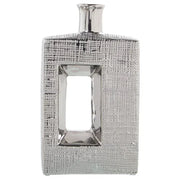 Vaso a bottiglia rettangolare verticale firmato Camacho, caratterizzato da un'elegante finitura ceramica color argento lucido con una texture a rilievo finemente lavorata e un originale design geometrico con foro centrale vuoto. Questo moderno complemento d'arredo è provvisto di un collo stretto superiore ideale per ospitare singoli steli floreali, perfetto come sofisticato oggetto ornamentale per impreziosire mobili, consolle o tavoli in contesti contemporanei.