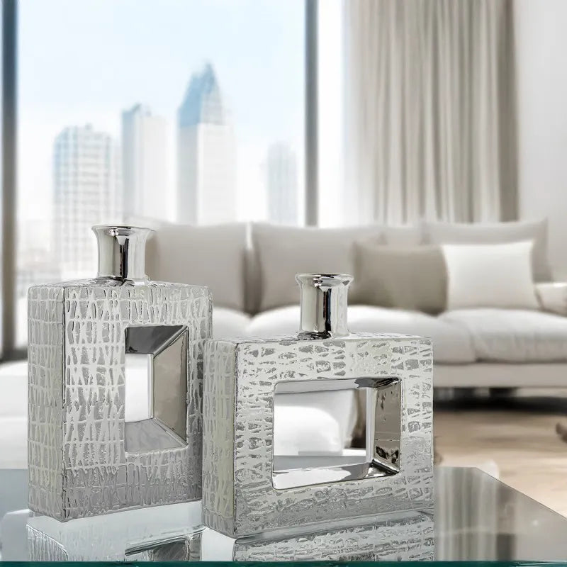 Vaso a bottiglia rettangolare orizzontale firmato Camacho, caratterizzato da un'elegante finitura ceramica color argento lucido con una texture a rilievo finemente lavorata e un originale design geometrico irregolare con foro centrale vuoto. Questo moderno complemento d'arredo è provvisto di un collo stretto superiore ideale per ospitare singoli steli floreali, perfetto come sofisticato oggetto ornamentale per impreziosire mobili, consolle o tavoli in contesti contemporanei.