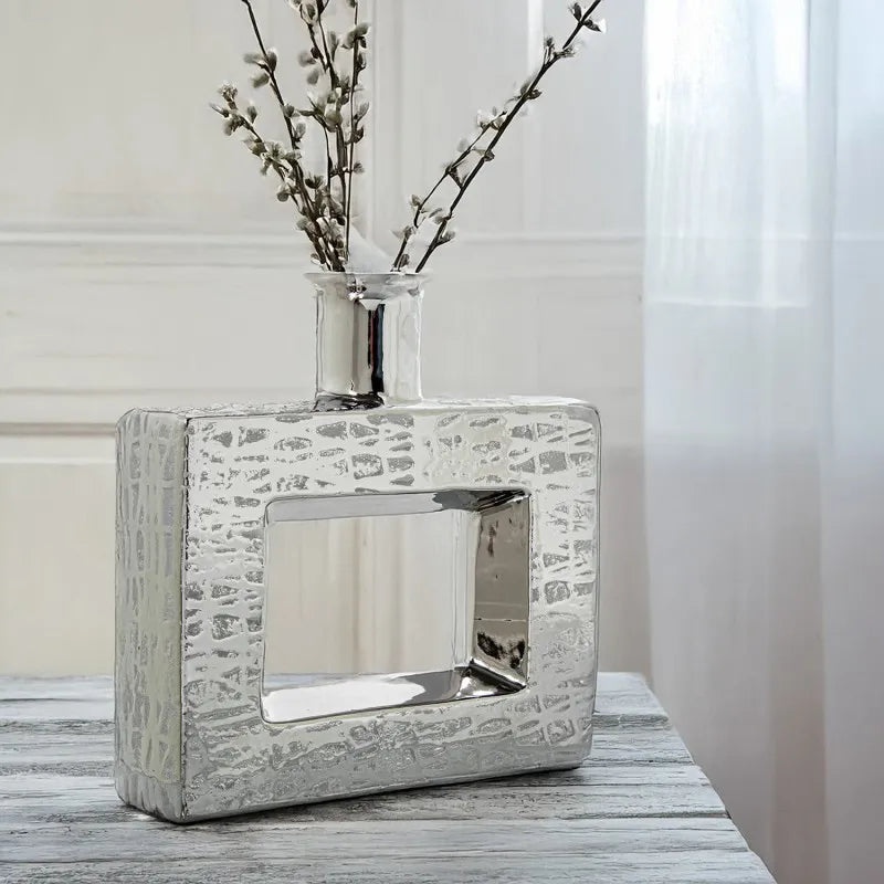 Vaso a bottiglia rettangolare orizzontale firmato Camacho, caratterizzato da un'elegante finitura ceramica color argento lucido con una texture a rilievo finemente lavorata e un originale design geometrico irregolare con foro centrale vuoto. Questo moderno complemento d'arredo è provvisto di un collo stretto superiore ideale per ospitare singoli steli floreali, perfetto come sofisticato oggetto ornamentale per impreziosire mobili, consolle o tavoli in contesti contemporanei.