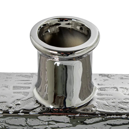 Vaso a bottiglia rettangolare orizzontale firmato Camacho, caratterizzato da un'elegante finitura ceramica color argento lucido con una texture a rilievo finemente lavorata e un originale design geometrico irregolare con foro centrale vuoto. Questo moderno complemento d'arredo è provvisto di un collo stretto superiore ideale per ospitare singoli steli floreali, perfetto come sofisticato oggetto ornamentale per impreziosire mobili, consolle o tavoli in contesti contemporanei.