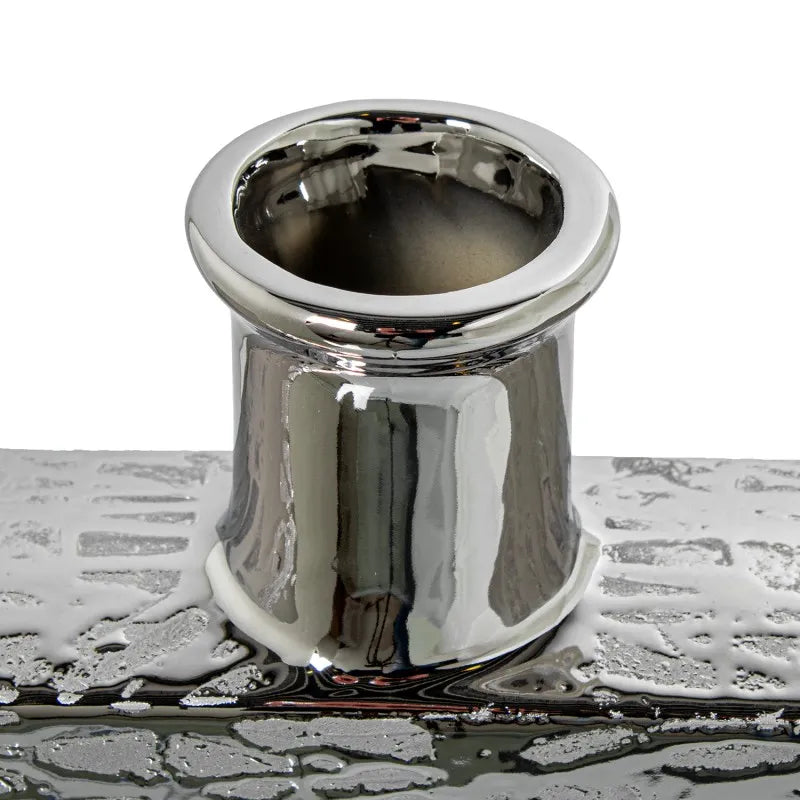 Vaso a bottiglia rettangolare orizzontale firmato Camacho, caratterizzato da un'elegante finitura ceramica color argento lucido con una texture a rilievo finemente lavorata e un originale design geometrico irregolare con foro centrale vuoto. Questo moderno complemento d'arredo è provvisto di un collo stretto superiore ideale per ospitare singoli steli floreali, perfetto come sofisticato oggetto ornamentale per impreziosire mobili, consolle o tavoli in contesti contemporanei.