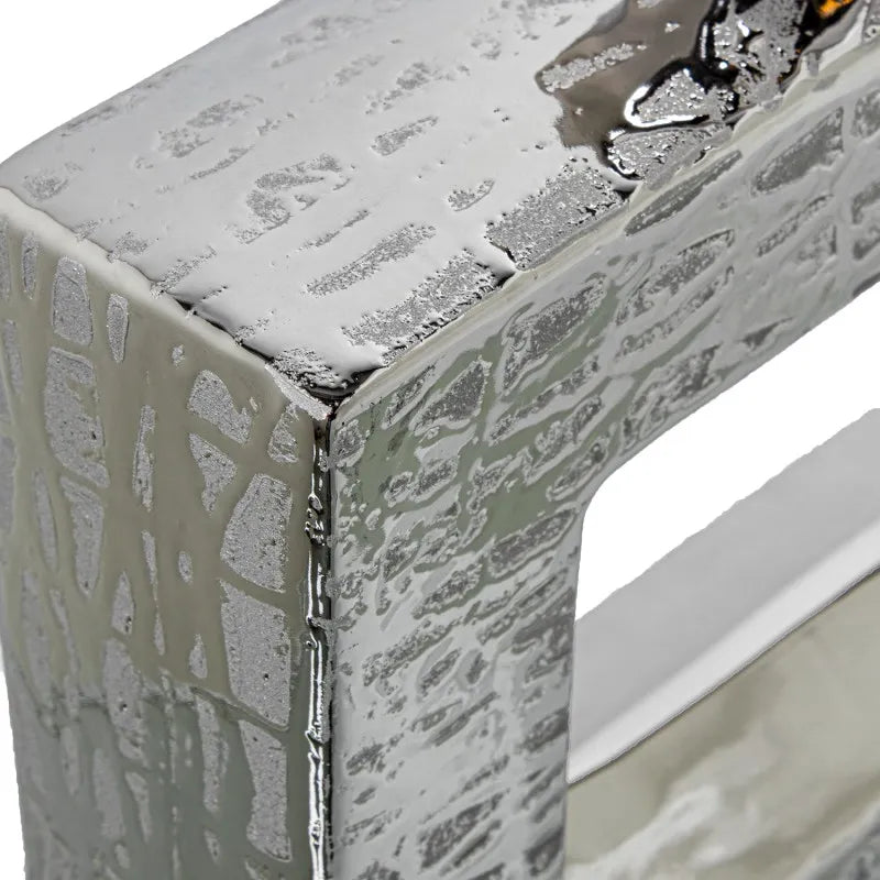 Vaso a bottiglia rettangolare orizzontale firmato Camacho, caratterizzato da un'elegante finitura ceramica color argento lucido con una texture a rilievo finemente lavorata e un originale design geometrico irregolare con foro centrale vuoto. Questo moderno complemento d'arredo è provvisto di un collo stretto superiore ideale per ospitare singoli steli floreali, perfetto come sofisticato oggetto ornamentale per impreziosire mobili, consolle o tavoli in contesti contemporanei.