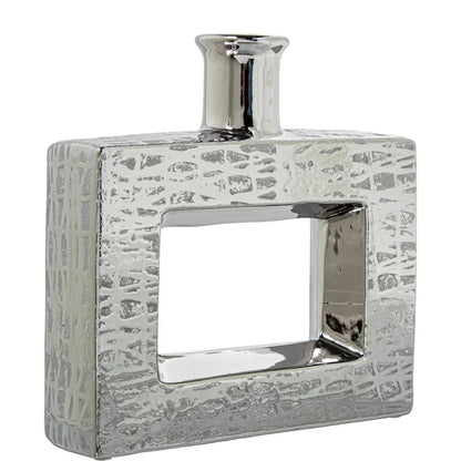 Vaso a bottiglia rettangolare orizzontale firmato Camacho, caratterizzato da un'elegante finitura ceramica color argento lucido con una texture a rilievo finemente lavorata e un originale design geometrico irregolare con foro centrale vuoto. Questo moderno complemento d'arredo è provvisto di un collo stretto superiore ideale per ospitare singoli steli floreali, perfetto come sofisticato oggetto ornamentale per impreziosire mobili, consolle o tavoli in contesti contemporanei.