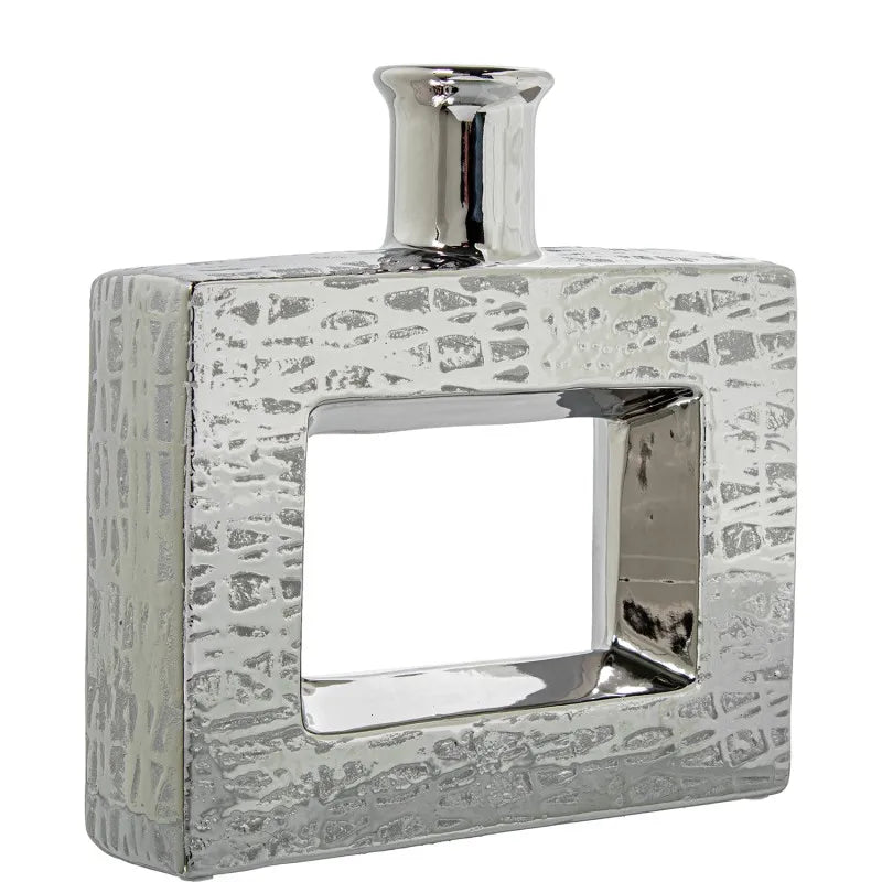 Vaso a bottiglia rettangolare orizzontale firmato Camacho, caratterizzato da un'elegante finitura ceramica color argento lucido con una texture a rilievo finemente lavorata e un originale design geometrico irregolare con foro centrale vuoto. Questo moderno complemento d'arredo è provvisto di un collo stretto superiore ideale per ospitare singoli steli floreali, perfetto come sofisticato oggetto ornamentale per impreziosire mobili, consolle o tavoli in contesti contemporanei.