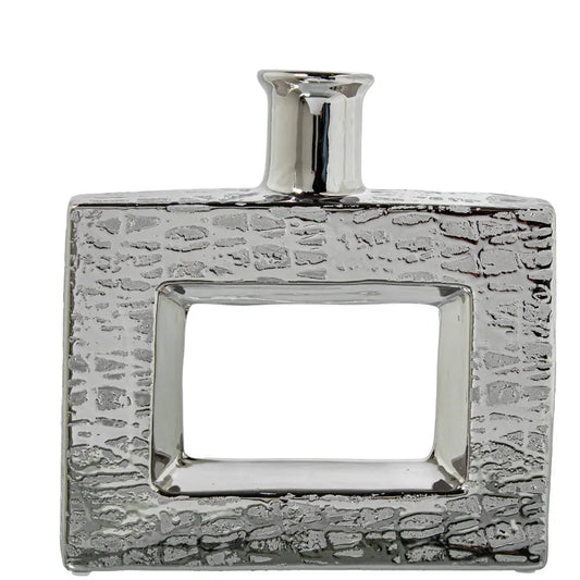 Vaso a bottiglia rettangolare orizzontale firmato Camacho, caratterizzato da un'elegante finitura ceramica color argento lucido con una texture a rilievo finemente lavorata e un originale design geometrico irregolare con foro centrale vuoto. Questo moderno complemento d'arredo è provvisto di un collo stretto superiore ideale per ospitare singoli steli floreali, perfetto come sofisticato oggetto ornamentale per impreziosire mobili, consolle o tavoli in contesti contemporanei.