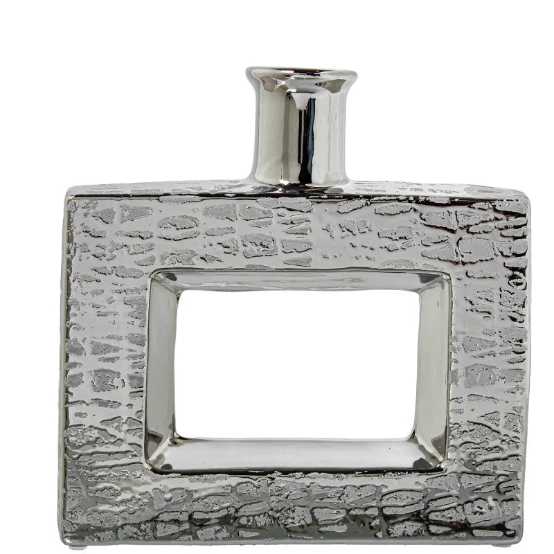 Vaso a bottiglia rettangolare orizzontale firmato Camacho, caratterizzato da un'elegante finitura ceramica color argento lucido con una texture a rilievo finemente lavorata e un originale design geometrico irregolare con foro centrale vuoto. Questo moderno complemento d'arredo è provvisto di un collo stretto superiore ideale per ospitare singoli steli floreali, perfetto come sofisticato oggetto ornamentale per impreziosire mobili, consolle o tavoli in contesti contemporanei.