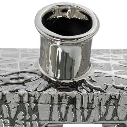 Vaso a bottiglia rettangolare verticale firmato Camacho, caratterizzato da un'elegante finitura ceramica color argento lucido con una texture a rilievo finemente lavorata e un originale design geometrico irregolare con foro centrale vuoto. Questo moderno complemento d'arredo è provvisto di un collo stretto superiore ideale per ospitare singoli steli floreali, perfetto come sofisticato oggetto ornamentale per impreziosire mobili, consolle o tavoli in contesti contemporanei.