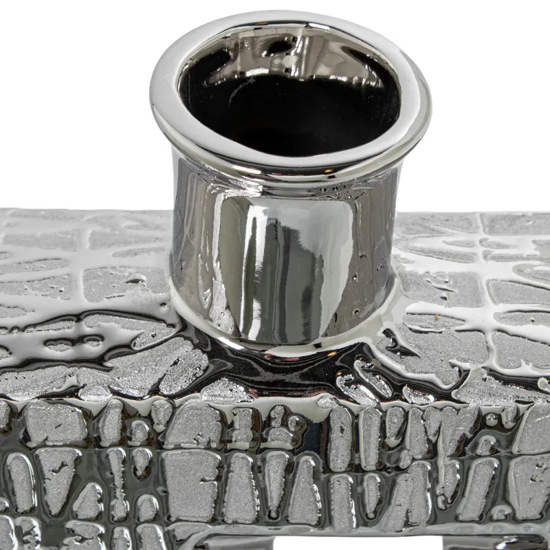 Vaso a bottiglia rettangolare verticale firmato Camacho, caratterizzato da un'elegante finitura ceramica color argento lucido con una texture a rilievo finemente lavorata e un originale design geometrico irregolare con foro centrale vuoto. Questo moderno complemento d'arredo è provvisto di un collo stretto superiore ideale per ospitare singoli steli floreali, perfetto come sofisticato oggetto ornamentale per impreziosire mobili, consolle o tavoli in contesti contemporanei.