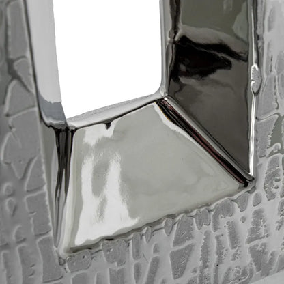 Vaso a bottiglia rettangolare verticale firmato Camacho, caratterizzato da un'elegante finitura ceramica color argento lucido con una texture a rilievo finemente lavorata e un originale design geometrico irregolare con foro centrale vuoto. Questo moderno complemento d'arredo è provvisto di un collo stretto superiore ideale per ospitare singoli steli floreali, perfetto come sofisticato oggetto ornamentale per impreziosire mobili, consolle o tavoli in contesti contemporanei.